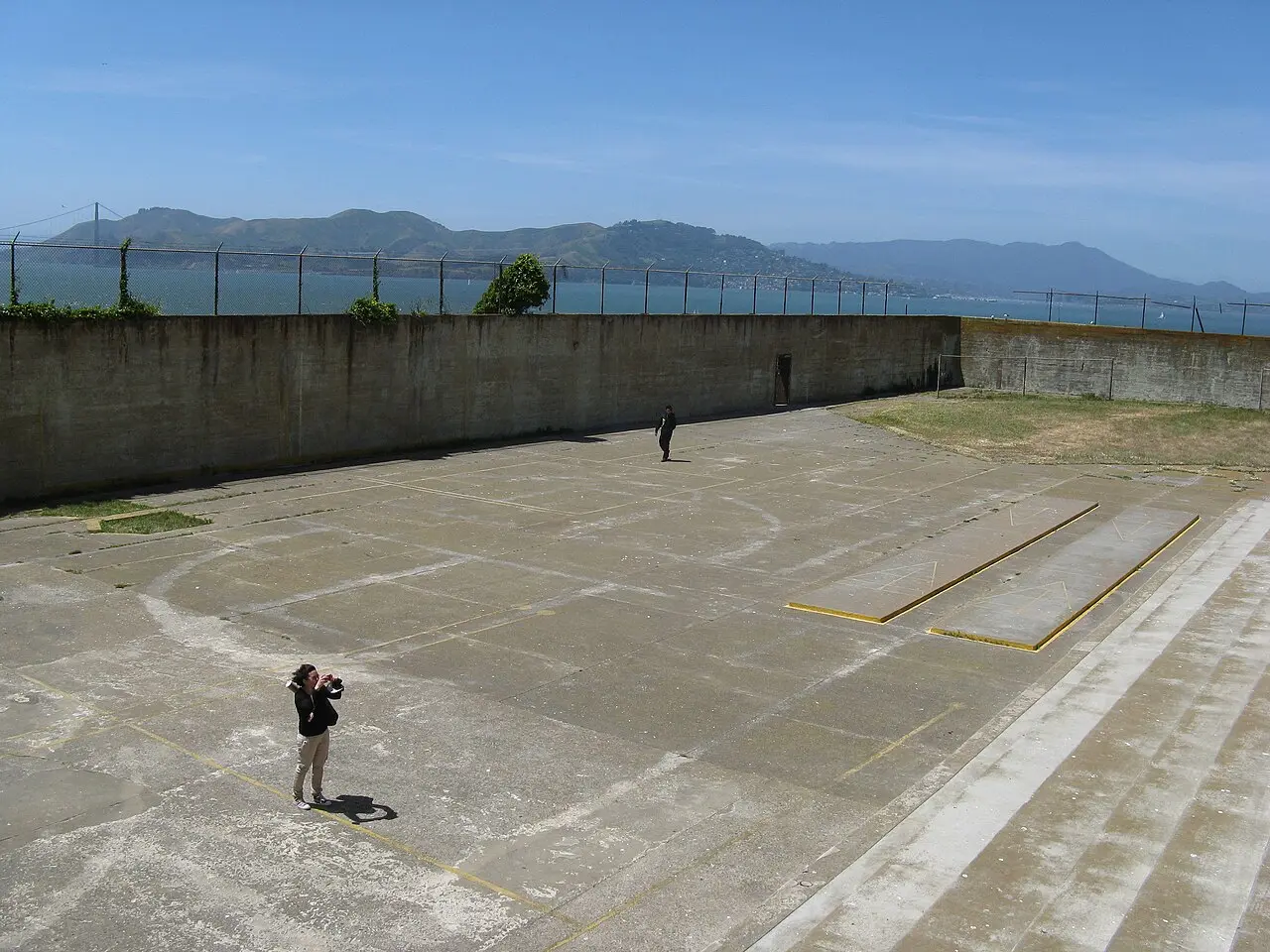 The concrete exercise yard at Alcatraz with a photographer and one other visitor, San Francisco Bay and hills visible beyond the wall