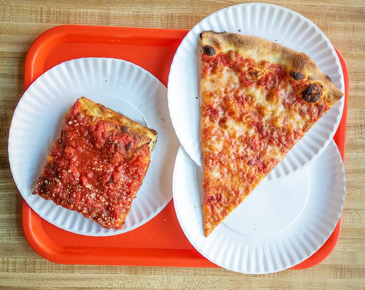 Two wide New York pizza slices on paper plates at a slice shop counter