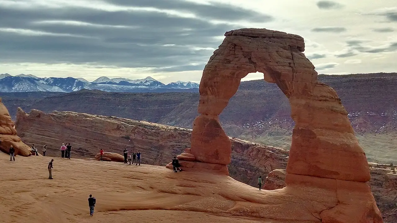 Delicate Arch freestanding over slickrock with hikers and snowy La Sal Mountains beyond