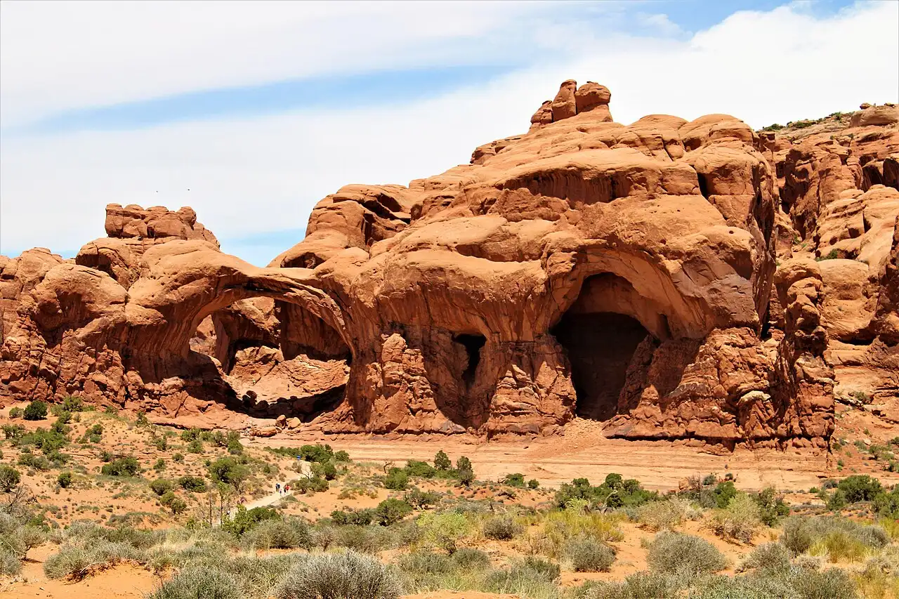 Massive Double Arch sandstone formation with two joined openings and tiny hikers on the sand below