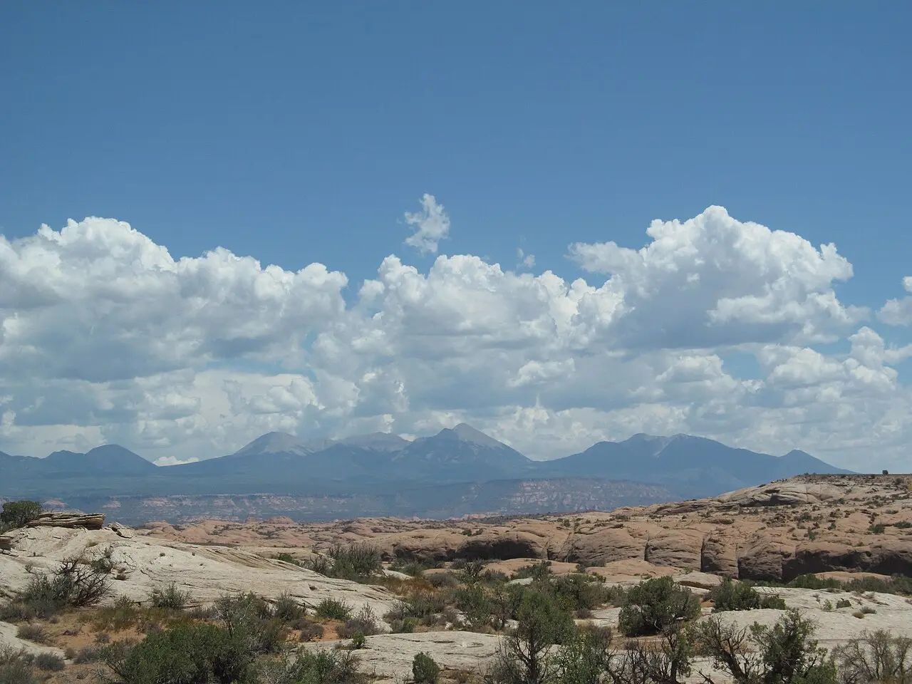 Slickrock desert foreground with distant mountain range and cumulus clouds under a blue Utah sky