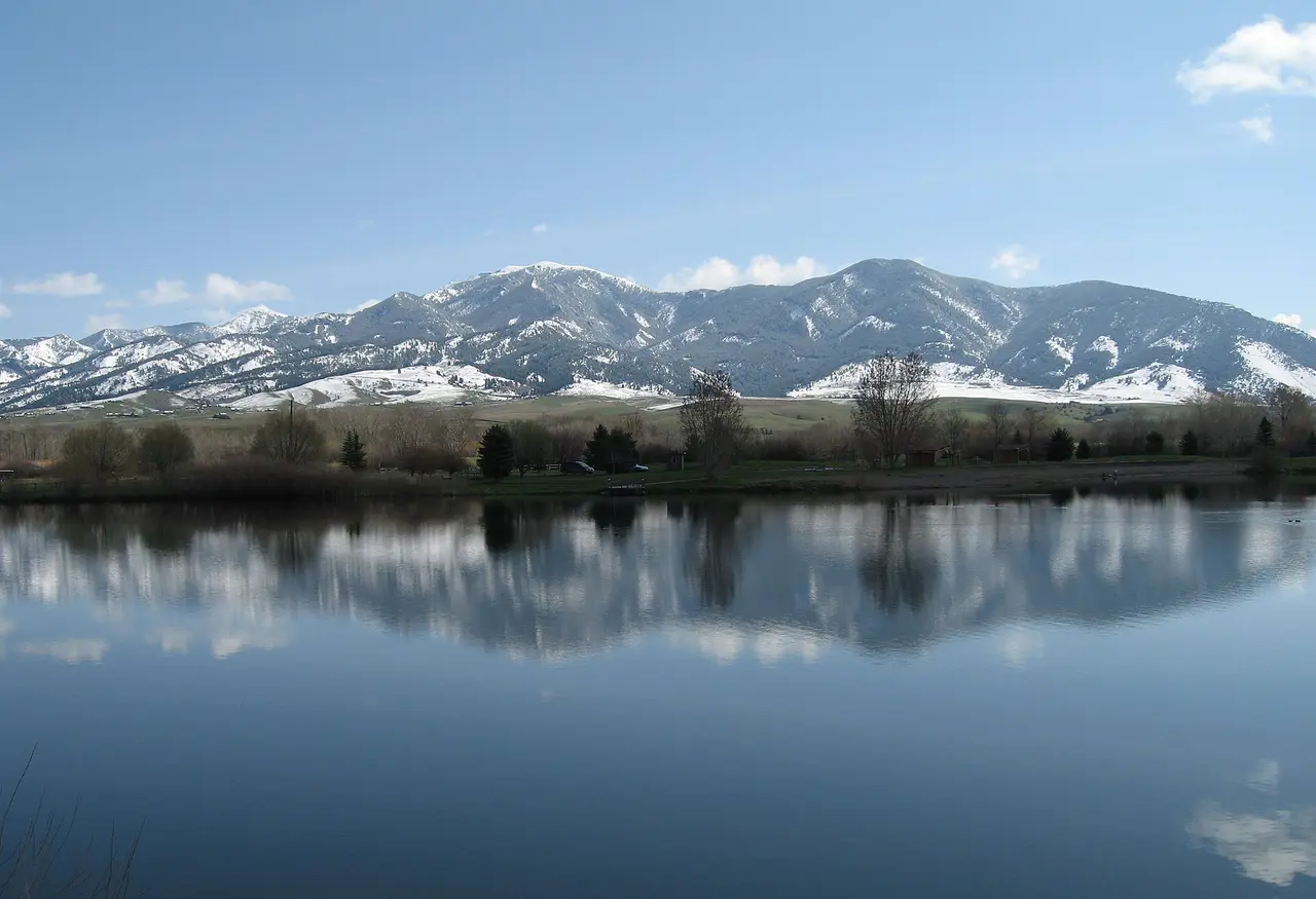 Still lake near Bozeman reflecting snow peaks, a thin shoreline of grass and trees