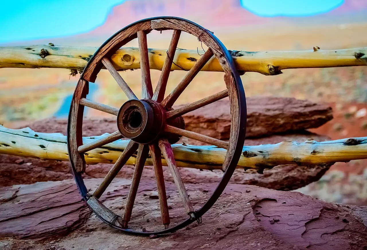 An old wooden wagon wheel leaning against a wooden fence with Monument Valley buttes visible in the background
