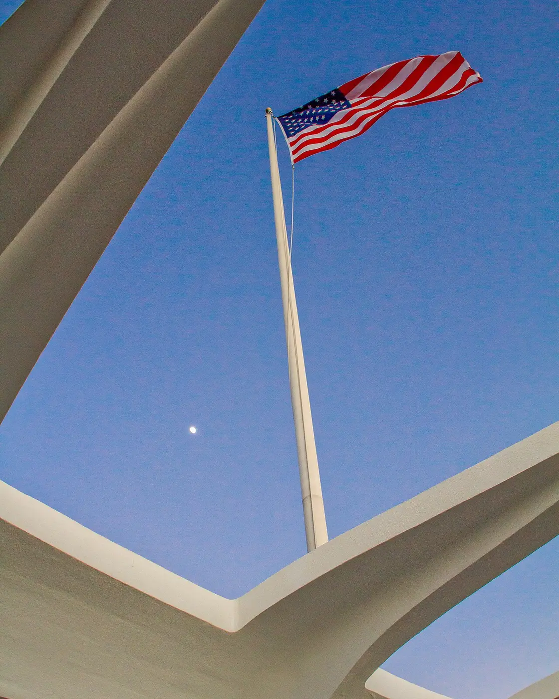American flag on a tall white pole framed by the open roof structure of the USS Arizona Memorial