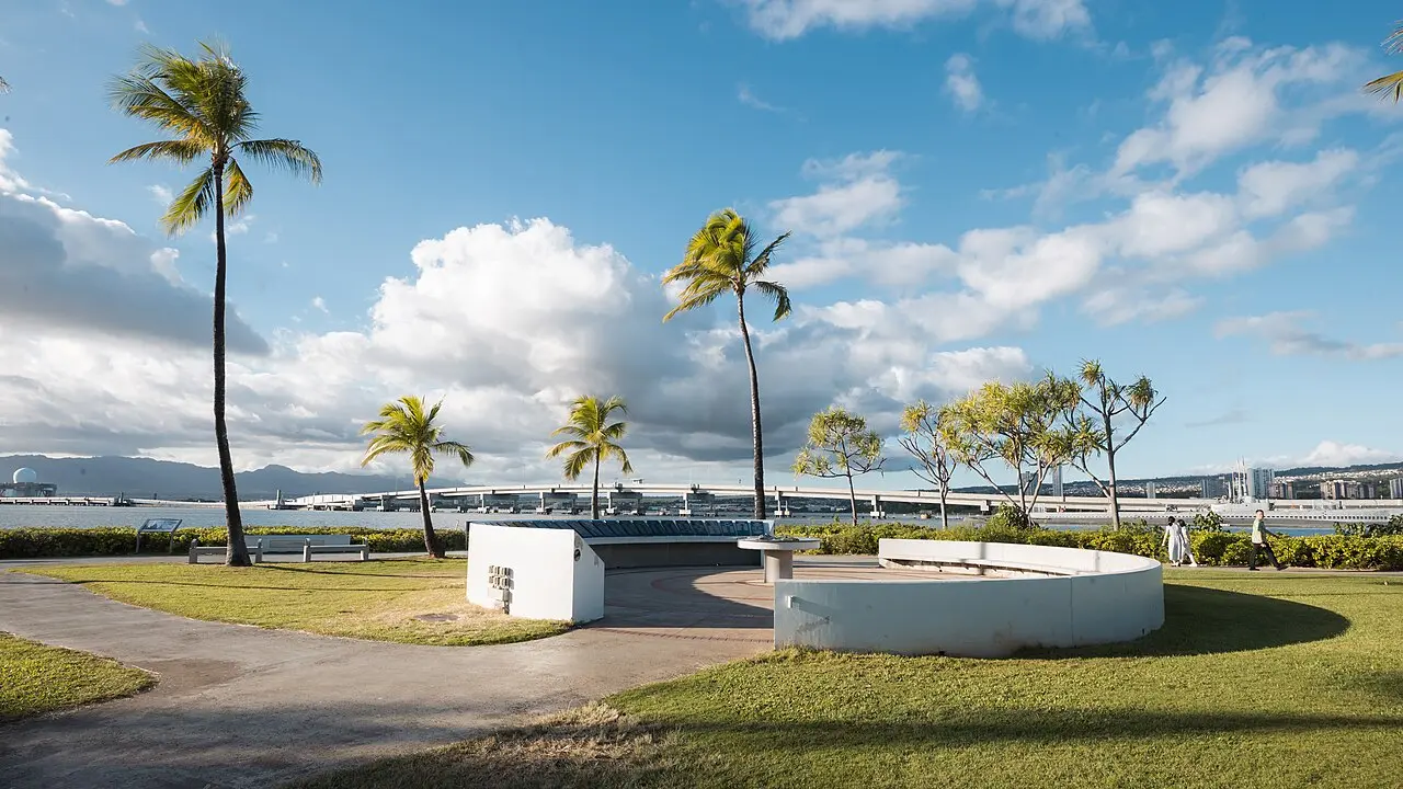 Palm trees and a circular memorial wall on the Pearl Harbor Visitor Center grounds with the harbor and Ford Island bridge beyond
