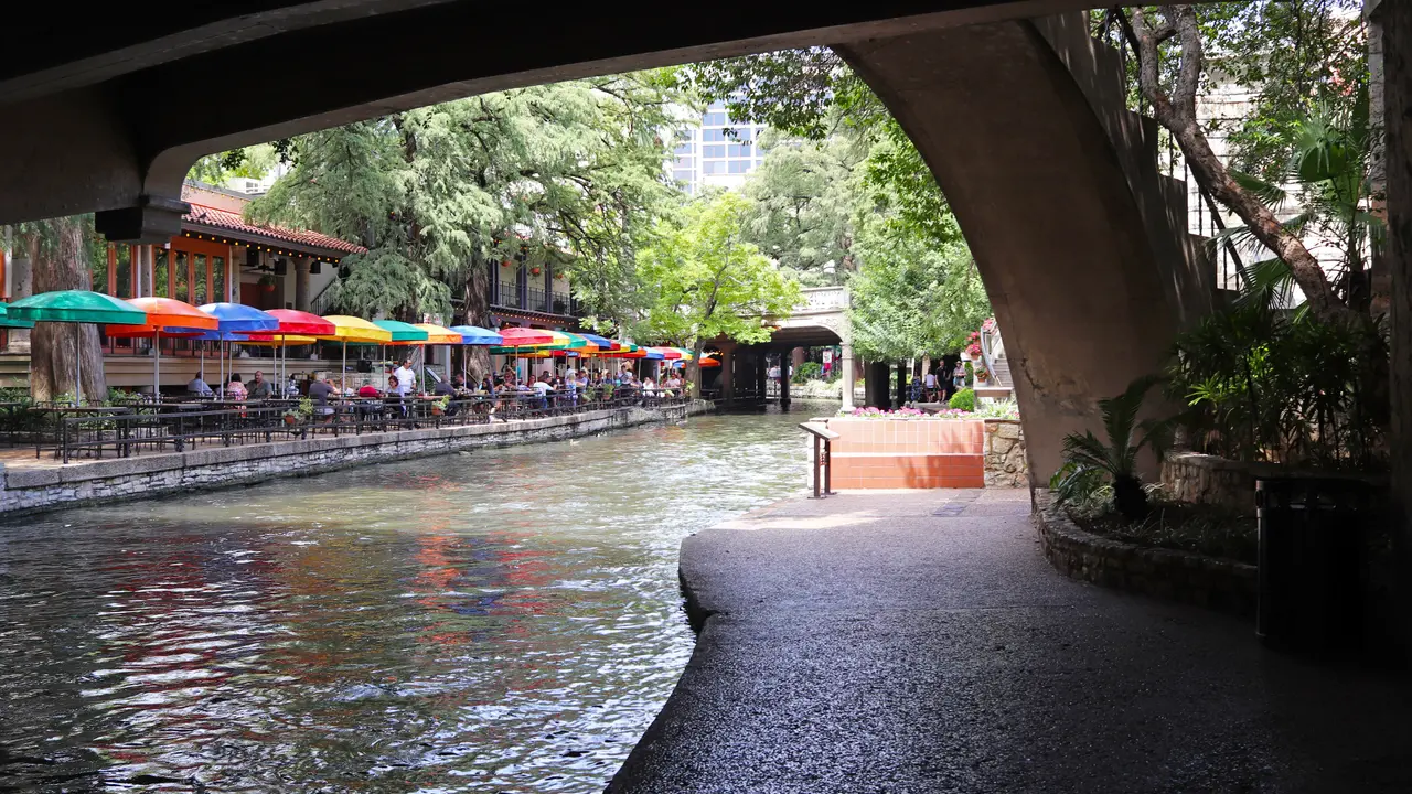 Colored umbrellas over restaurant terraces along the San Antonio River Walk seen from under a bridge
