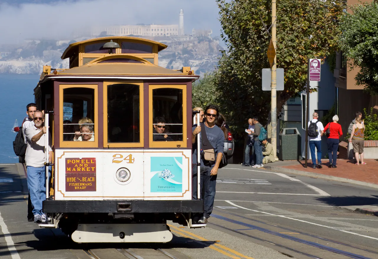 A cable car climbing Hyde Street with Alcatraz Island visible in the bay behind