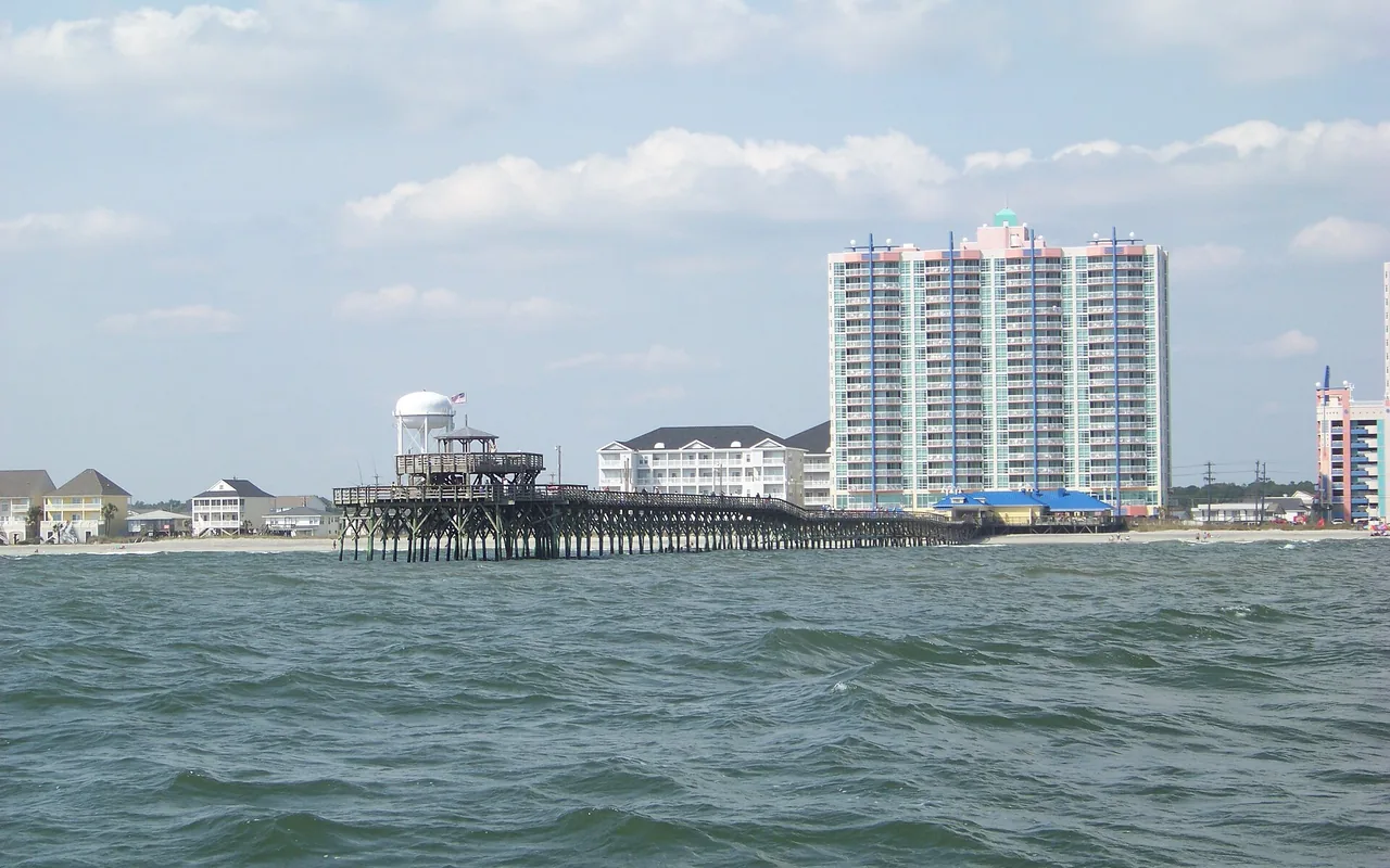 A fishing pier reaching into the ocean with beachfront condo towers and low beach houses behind it on the Grand Strand