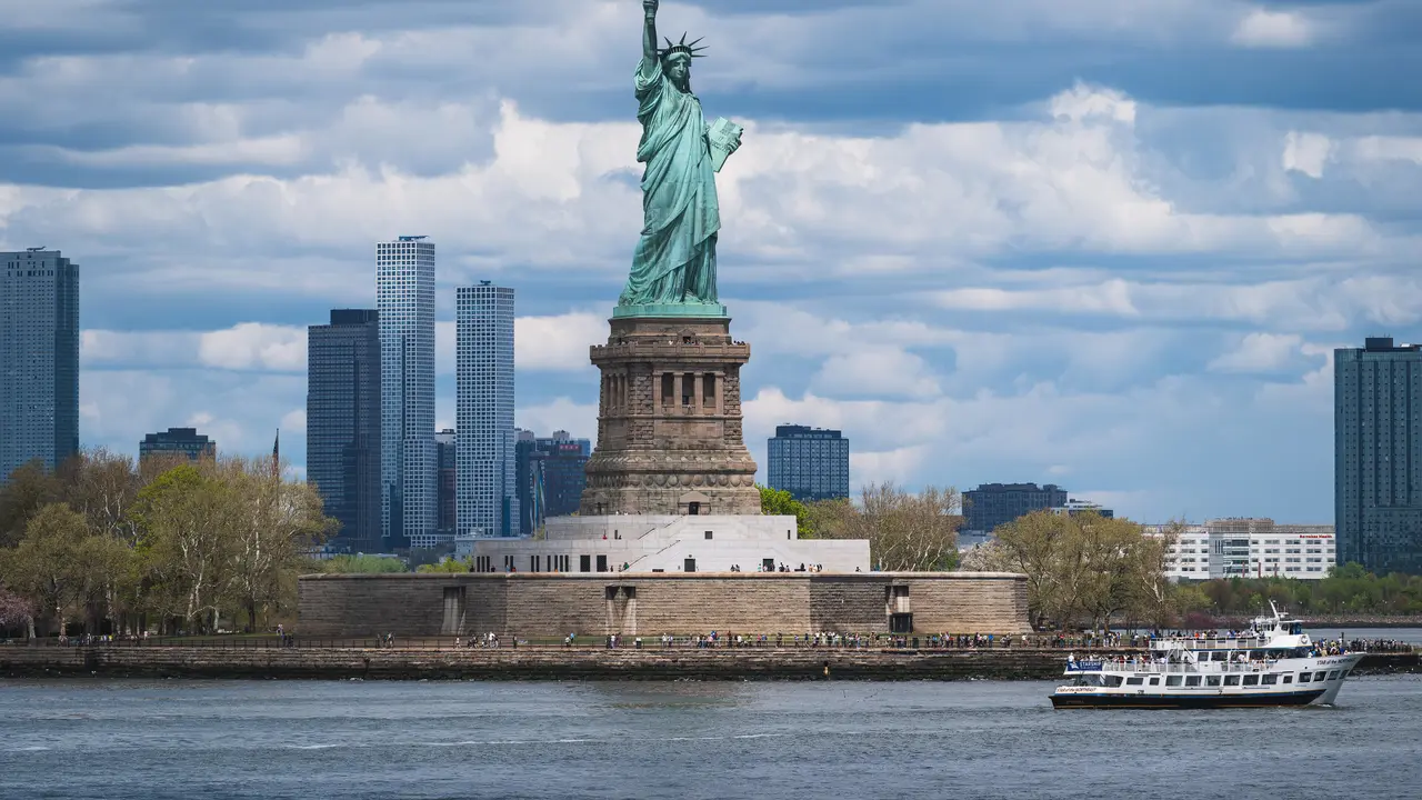 The Statue of Liberty on Liberty Island with a ferry passing in front and the Jersey City skyline behind