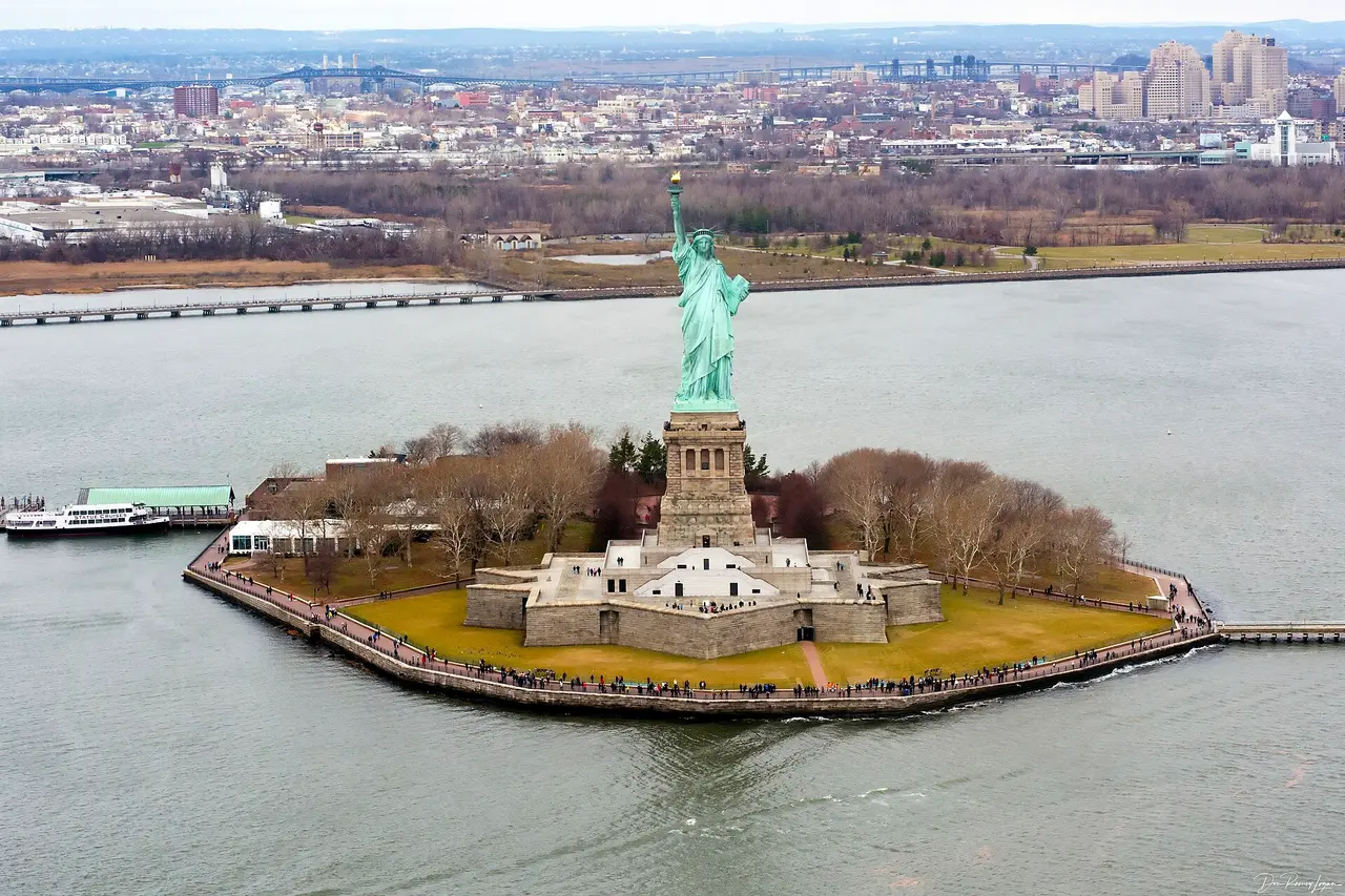 Liberty Island seen from the air with the ferry dock, the star-shaped fort base and visitors on the perimeter path